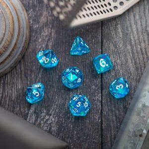 May include: A set of seven translucent blue polyhedral dice for tabletop games. Each die has white numbering and a faceted design. The dice are arranged on a dark wooden surface.