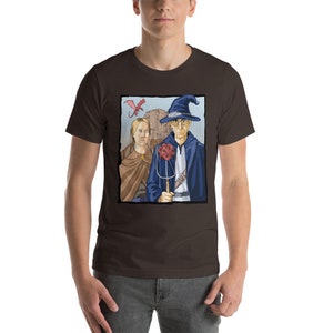 May include: A brown t-shirt with a graphic of a wizard and a woman standing in front of a castle. A red dragon flies above them. The wizard is holding a staff with a red twenty-sided die on top.