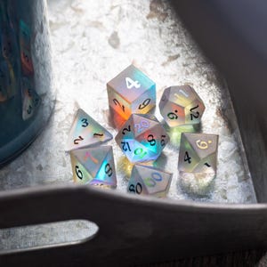 May include: A set of iridescent dice with black numbers on each face. The dice are made of clear glass and have a rainbow effect. The dice are on a metal tray.