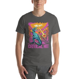 May include: A dark gray t-shirt with a graphic of a blue dinosaur standing on a pink and orange landscape with falling stars. The text "CRITICAL HIT" is printed in pink below the dinosaur.