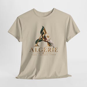 Könnte beinhalten: Ein beiges Kurzarm-T-Shirt mit einem großen goldenen Buchstaben A, der mit einem Halbmond und einem Stern verziert ist. Unter dem Buchstaben steht das Wort ALGÉRIE in Gold, gefolgt von dem Text FIERTÉ • RÉSILIENCE • ESPOIR. Das Design enthält grüne, weiße und rote Akzente.