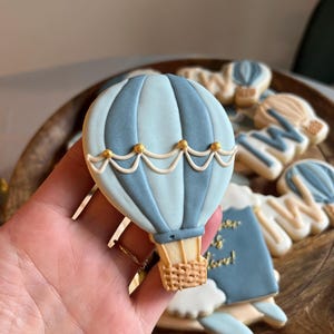 May include: A blue and white hot air balloon cookie decorated with gold accents. The cookie is held in a hand and is surrounded by other cookies.