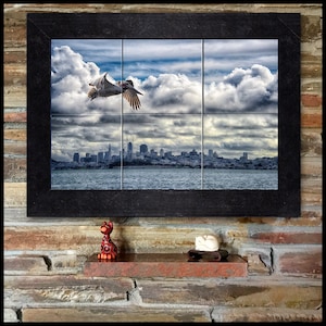 Puede incluir: Un mosaico de azulejos enmarcado que representa un pelícano blanco volando sobre una ciudad con un cielo nublado. La ciudad es una vista lejana de una ciudad con edificios altos y una masa de agua en primer plano.