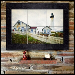 Puede incluir: Una pintura enmarcada que representa un faro blanco con una valla blanca de madera delante. El faro está en una colina con vistas al mar. Hay dos edificios blancos en primer plano, uno con un techo rojo y el otro con un techo azul.