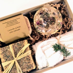 Puede incluir: Una caja de regalo llena de productos de baño, incluyendo dos barras de jabón, una bomba de baño y una toalla blanca. Las barras de jabón están etiquetadas con "Lavender & Rose with Oatmeal".