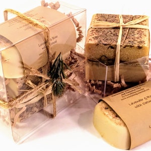 Puede incluir: Tres cajas de plástico transparente llenas de papel triturado y atadas con cuerda. Cada caja contiene una barra de jabón artesanal envuelta en papel marrón con el texto "Lavender & Rose with Oatmeal".