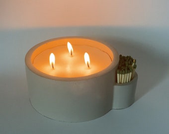 May include: A white concrete candle holder with three lit candles and a side compartment for matches. The matches are green and white.