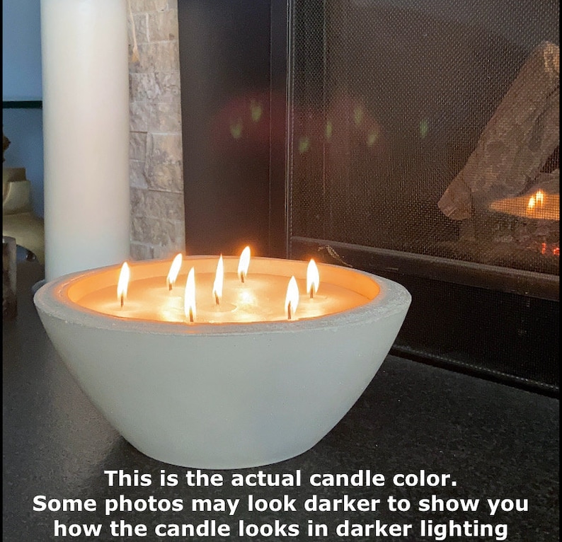 May include: A white concrete bowl candle with multiple wicks burning. The candle is lit and the flames are visible. The candle is sitting on a dark surface.