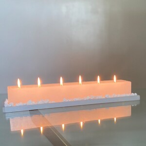 Puede incluir: Una vela rectangular de color rosa claro con seis mechas encendidas. La vela se asienta sobre una base rectangular blanca, decorada con peque&ntilde;as piedras blancas en forma de cristal. La vela y la base se reflejan en una superficie de vidrio.