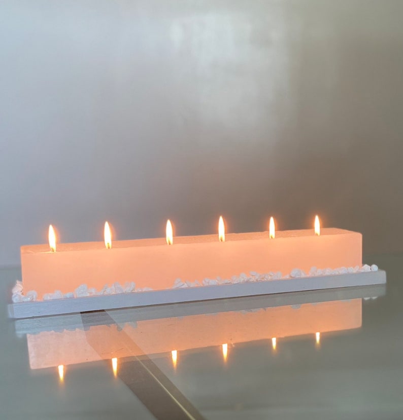 Puede incluir: Una vela rectangular de color rosa claro con seis mechas encendidas. La vela se asienta sobre una base rectangular blanca, decorada con peque&ntilde;as piedras blancas en forma de cristal. La vela y la base se reflejan en una superficie de vidrio.