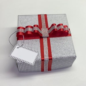 May include: A silver gift box with a red ribbon and bow. The box is covered in small silver sequins and has a white gift tag attached.