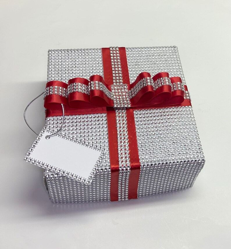 May include: A silver gift box with a red ribbon and bow. The box is covered in small silver sequins and has a white gift tag attached.