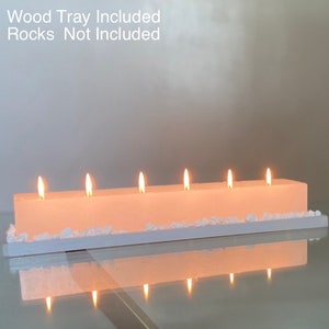 Puede incluir: Un portavelas rectangular blanquecino con seis velas encendidas. Las velas est&aacute;n sobre una bandeja blanca, con piedras decorativas blancas. El texto "Wood Tray Included Rocks Not Included" es visible.