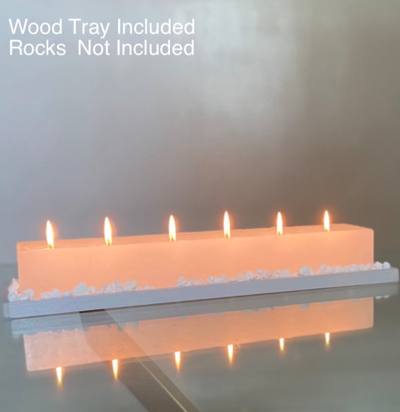 Puede incluir: Un portavelas rectangular blanquecino con seis velas encendidas. Las velas est&aacute;n sobre una bandeja blanca, con piedras decorativas blancas. El texto "Wood Tray Included Rocks Not Included" es visible.