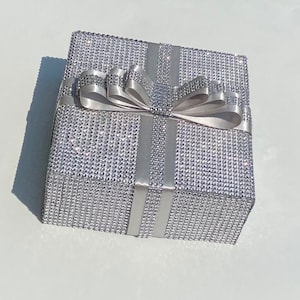 May include: A silver gift box with a large silver bow. The box is covered in small, sparkling rhinestones.