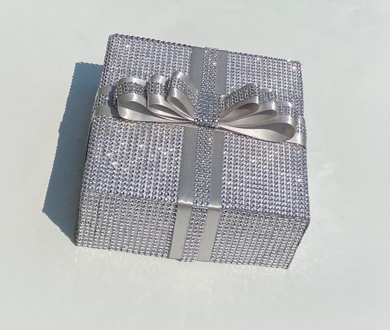 May include: A silver gift box with a large silver bow. The box is covered in small, sparkling rhinestones.