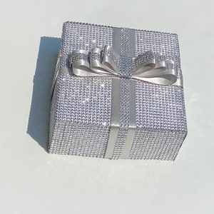 May include: A silver gift box with a silver ribbon and bow. The box is covered in small, sparkly rhinestones.