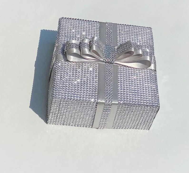 May include: A silver gift box with a silver ribbon and bow. The box is covered in small, sparkly rhinestones.