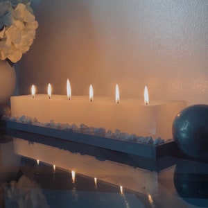 Puede incluir: Una vela blanca rectangular grande con seis mechas encendidas. La vela se encuentra sobre una bandeja de madera con peque&ntilde;as piedras blancas. Un arreglo floral blanco y una esfera plateada est&aacute;n en el fondo.