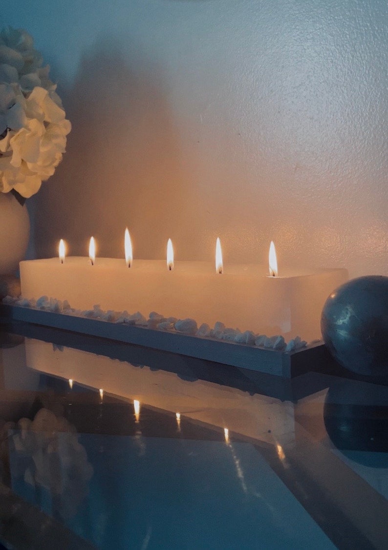 Puede incluir: Una vela blanca rectangular grande con seis mechas encendidas. La vela se encuentra sobre una bandeja de madera con peque&ntilde;as piedras blancas. Un arreglo floral blanco y una esfera plateada est&aacute;n en el fondo.