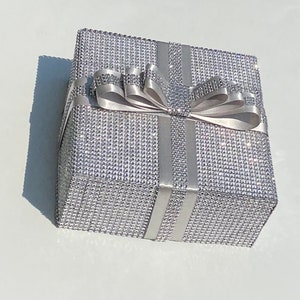 May include: A silver gift box with a large white satin bow. The box is covered in small, sparkling rhinestones.