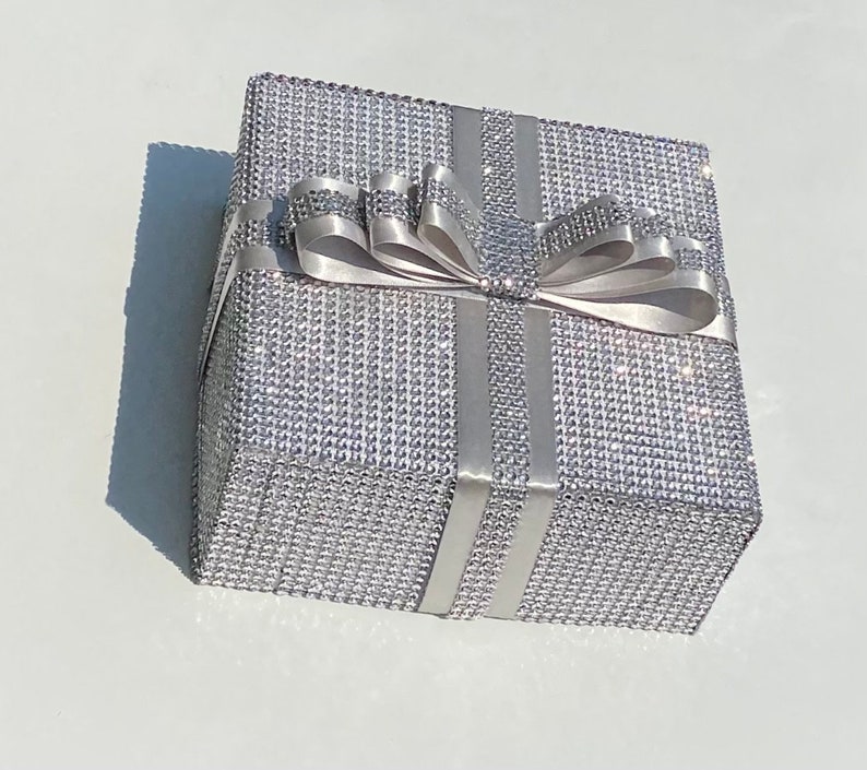 May include: A silver gift box with a large white satin bow. The box is covered in small, sparkling rhinestones.