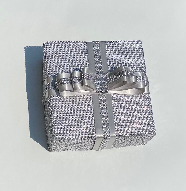 May include: A silver gift box with a sparkly, textured surface and a large silver bow. The box is tied with silver ribbon.
