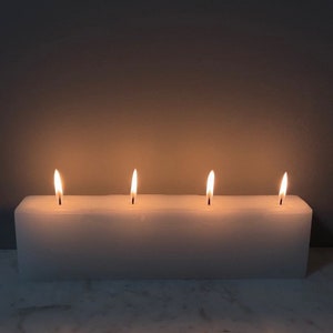May include: A white rectangular candle with three wicks burning. The candle is sitting on a white surface.