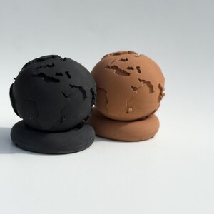 May include: Two ceramic globe sculptures, one black and one terracotta, each with continents carved out. The globes sit atop matching circular bases. The background is plain white.