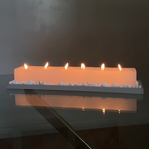 Puede incluir: Una vela rectangular de color claro con seis llamas encendidas. La vela est&aacute; sobre una bandeja gris, rodeada de peque&ntilde;as piedras blancas. El reflejo de la vela y las llamas es visible en la superficie de vidrio de abajo.