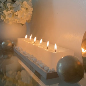 Puede incluir: Un portavelas rectangular blanco con seis velas encendidas. Las velas est&aacute;n colocadas en una base blanca con peque&ntilde;as piedras blancas. Un jarr&oacute;n blanco con flores blancas y esferas doradas se encuentra en el fondo.