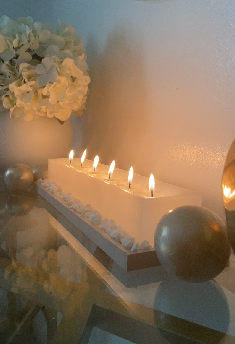 Puede incluir: Un portavelas rectangular blanco con seis velas encendidas. Las velas est&aacute;n colocadas en una base blanca con peque&ntilde;as piedras blancas. Un jarr&oacute;n blanco con flores blancas y esferas doradas se encuentra en el fondo.