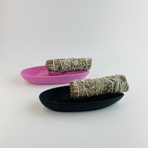 May include: Two oval-shaped incense holders, one black and one pink, each holding a bundle of white sage. The sage bundles are wrapped with white string. The holders are on a white background.