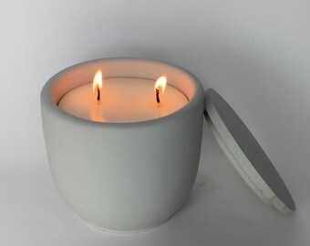 May include: A gray concrete candle jar with a lid, the candle is lit with two wicks.