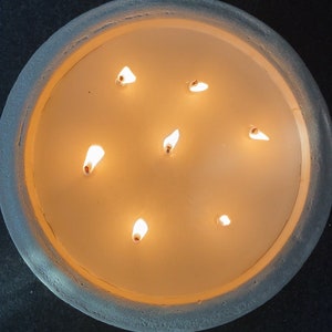 May include: A round, white candle holder with a textured surface. The candle holder has seven lit candles inside.