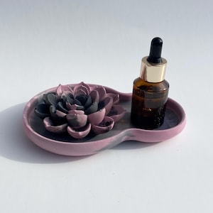 May include: A pink and gray concrete tray with a black and pink flower design. A small brown glass bottle with a gold lid and a black dropper sits on the tray.