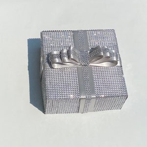 May include: A silver gift box with a sparkly, textured surface and a white satin ribbon tied in a bow.