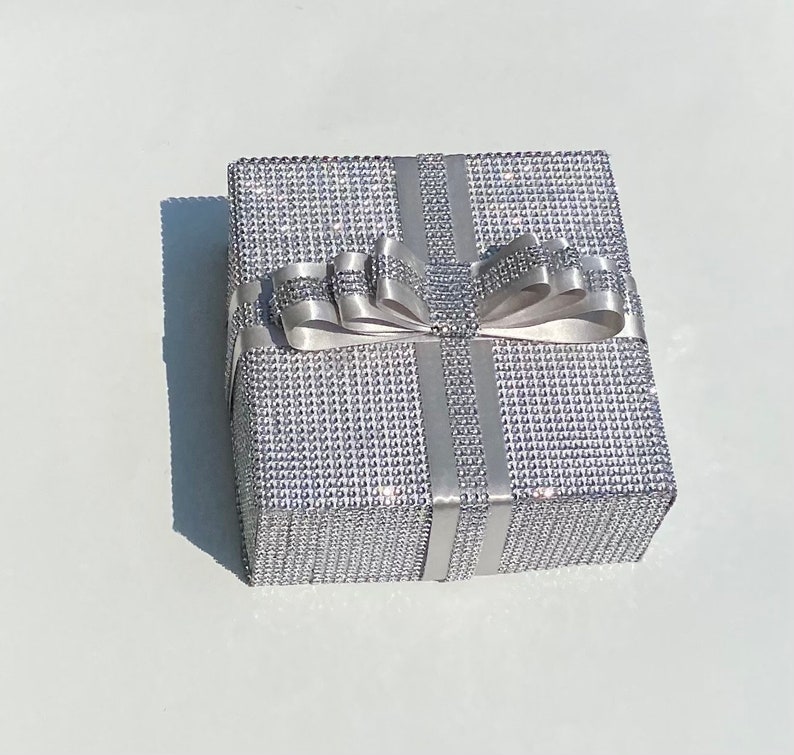 May include: A silver gift box with a sparkly, textured surface and a white satin ribbon tied in a bow.