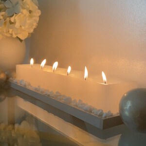 Puede incluir: Una vela blanca rectangular con seis mechas encendidas. La vela se asienta sobre una base de madera blanca, decorada con peque&ntilde;as piedras blancas. Un jarr&oacute;n blanco con flores blancas y una esfera decorativa est&aacute;n en el fondo.