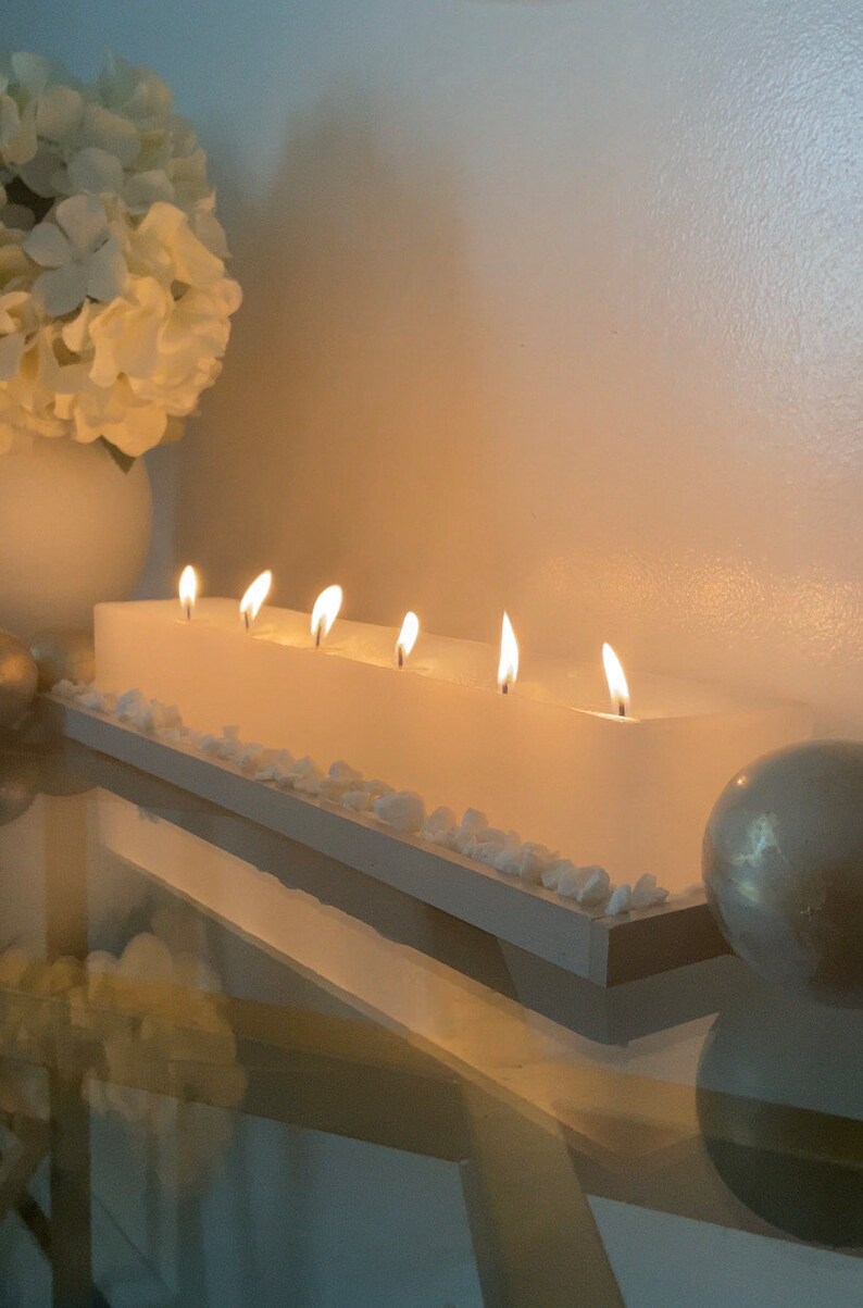 Puede incluir: Una vela blanca rectangular con seis mechas encendidas. La vela se asienta sobre una base de madera blanca, decorada con peque&ntilde;as piedras blancas. Un jarr&oacute;n blanco con flores blancas y una esfera decorativa est&aacute;n en el fondo.