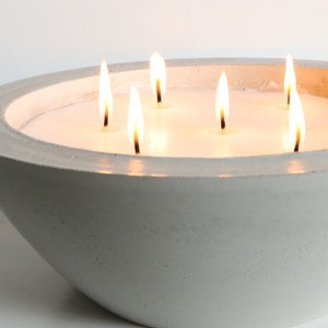 May include: A white concrete bowl candle with six wicks burning. The candle is lit and the flames are visible.