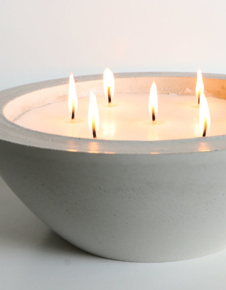 May include: A white concrete bowl candle with six wicks burning. The candle is lit and the flames are visible.