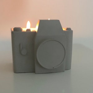 May include: A gray concrete candle shaped like a vintage camera. The candle has a burning wick, emitting a warm glow. The camera design includes a lens, buttons, and a flash. A unique decorative item.