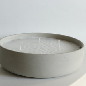 May include: A large, round, gray concrete candle with multiple wicks. The candle wax is white and fills the bowl-shaped container. The candle is a decorative item for home decor.