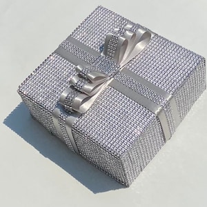 May include: A silver gift box with a sparkly, textured surface and a white satin ribbon tied in a bow.
