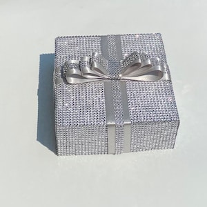 May include: A silver gift box with a large silver bow. The box is covered in small, sparkling rhinestones.