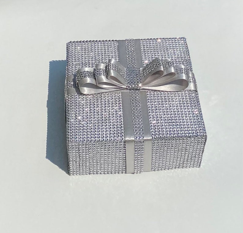 May include: A silver gift box with a large silver bow. The box is covered in small, sparkling rhinestones.