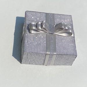 May include: A silver gift box with a sparkly, diamond-like finish. The box is tied with a silver satin ribbon and bow.