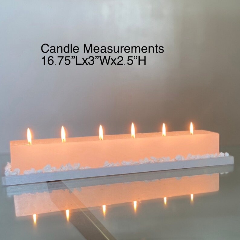 Puede incluir: Una vela rectangular con seis mechas encendidas. La vela es de color rosa claro y descansa sobre una base blanca con peque&ntilde;as piedras blancas. El texto "Candle Measurements 42.5cmLx7.6cmWx6.4cmH" es visible.