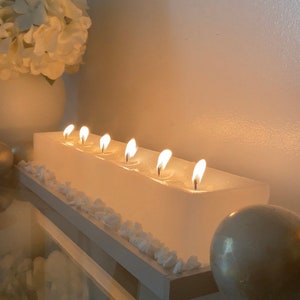 Puede incluir: Un portavelas rectangular blanco con siete velas encendidas. Las velas est&aacute;n colocadas en una base blanca, decorada con peque&ntilde;as piedras blancas. Un jarr&oacute;n blanco con flores blancas se encuentra en el fondo.